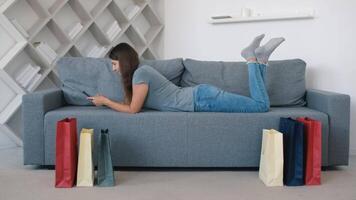 Online shopping, Buying from home, Credit card, Online store. Beautiful woman chooses a product in an online store lying on the couch at home using a smartphone video