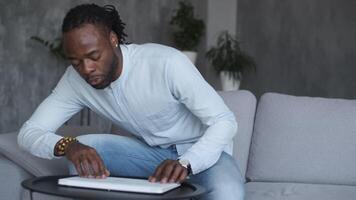 A male African American freelancer sits down on a sofa, turns on a laptop and starts an online webinar. video