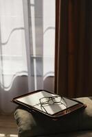 Glasses and an open weekly book lay on a couch in a room with dimmed light. There is a beautiful pattern of light and shadow on the furniture and on the diary. photo