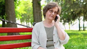 idosos mulher sentado em parque Banco leva telefone chamar. ativo aposentadoria estilo de vida e gastos Tempo ao ar livre video