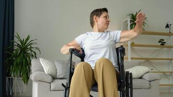 Mature 50 yers woman is spreading her arms in different sides in wheelchair. Training, sport, recovery and lifting, stretching exercise at home video