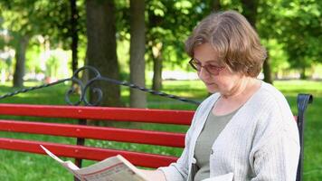 Senior woman in glasses reading newspaper in park. Retirement age video