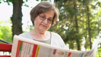 senior vrouw lezing krant, zittend Aan bank in park. pensioen leeftijd video