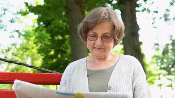 senior vrouw lezing krant, zittend Aan bank in park. pensioen leeftijd video