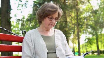 Relaxed senior woman reads book sitting on bench in park. Active retirement lifestyle, hobby and spending time outdoors video