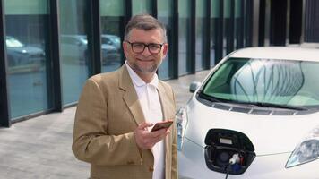 retrato de más viejo hombre con teléfono inteligente mientras cargando eléctrico coche. cargando cable atascado en a cero emisión eléctrico coche video