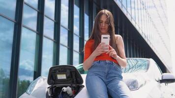 Attractive woman uses phone while charging electric car near the shopping center video