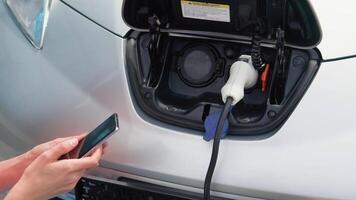 Woman puts charger into her electric car and use phone when waiting. Woman plugging in power cable to zero emission electric car at charging station with city landscape video