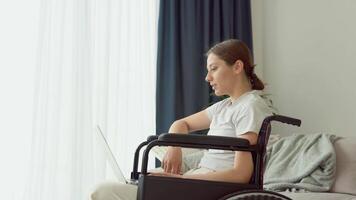 Smiling young woman in wheelchair chatting on social networks at home. Happy girl web surfing information video