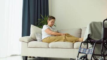 Senior woman massaging her legs while sitting on the couch near the wheelchair video