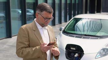 hermoso más viejo empresario utilizando teléfono inteligente mientras cargando eléctrico coche. cargando cable atascado en a cero emisión eléctrico coche video