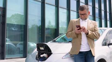 Sénior homme parle sur le téléphone tandis que le électrique voiture est mise en charge en plein air. prise de courant mise en charge un électrique voiture de mise en charge station video