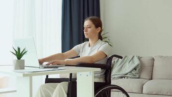 Female student learning online using laptop typing sitting in wheelchair in apartment busy with distance education video