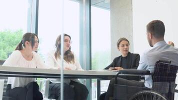 diverse groupe de travail collègues parlant à Bureau réunion, un dans fauteuil roulant. indépendant Créatif affaires dans une moderne Bureau video