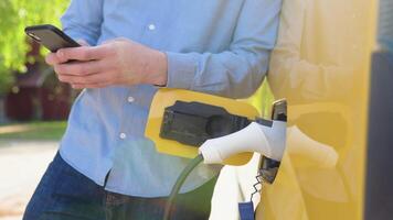 joven hombre charla en móvil teléfono mientras eléctrico coche es cargando en al aire libre estación video