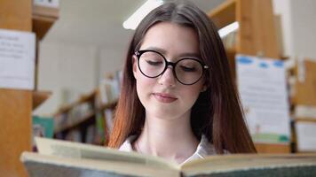 un' sorridente giovane ragazza nel bicchieri legge un' libro nel il Università biblioteca. un' riflessivo giovane alunno legge un' libro in piedi nel il biblioteca video