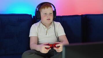 een jongen gamer is zittend Aan een bankstel in voorkant van de monitor, spelen en winnend in spellen Aan troosten. knus kamer met warm en neon licht video