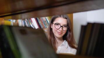 ein nachdenklich jung Schüler liest ein Buch Stehen im das Bibliothek. ein lächelnd jung Mädchen im Brille liest ein Buch im das Universität Bibliothek video