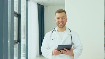 Successful physician in white lab coat with tablet looks at the camera and smiles in hospital office video