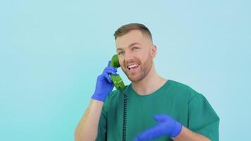 A friendly doctor in a green uniform and blue gloves speaks on an old telephone and consults a patient video