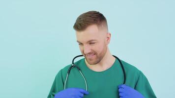 exitoso médico cirujano en verde uniforme, azul guantes y estetoscopio mira a cámara y sonrisas en azul antecedentes video