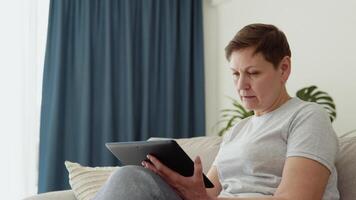 Smiling elderly mature woman resting on sofa, using digital tablet alone at home. Happy older pensioner web surfing information. Studying or shopping online video