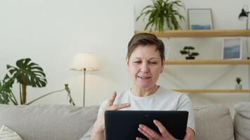 Smiling elderly mature woman resting on sofa, using digital tablet alone at home. Chatting on social networks video