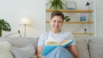 Relaxed senior woman sitting on sofa and reading book indoors at home. Leisure activity of mature female at comfort modern flat. Casual elderly life and wrinkled skin closeup video