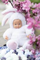 Baby doll in a white Easter bunny costume, against a background of sakura flowers and Easter eggs, Easter concept photo
