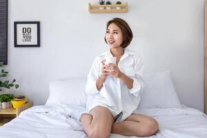 Young pretty girl holding cup of coffee on her bed looking outside the window for morning routine usage photo