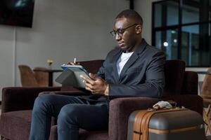 africano americano empresario es utilizando digital tableta mientras esperando en aerolínea negocio salida salón esperando para embarque avión foto