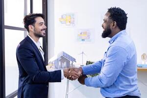 Caucasian business architect congratulate his African engineer for outstanding achievement team performance by shaking hand in modern real estate office workplace with floor plan and layout photo