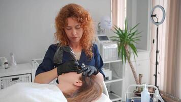 Aesthetic cosmetology. Master makes permanent eyebrow makeup procedure using special needle tattoo machine to woman in beauty salon. video
