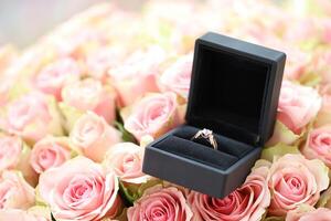 Gold diamond engagement ring in black box case among big amount of roses in big bouquet close up photo