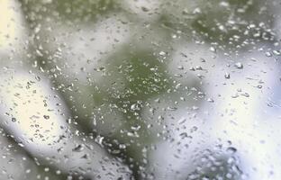 A photo of rain drops on the window glass with a blurred view of the blossoming green trees. Abstract image showing cloudy and rainy weather conditions