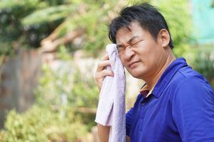 Asian man feel headache and unwell from hot weather condition, use cool wet cloth to wipe face for relief symptom. Concept, self first aid to protect from heat stroke, cool down. Health care. photo