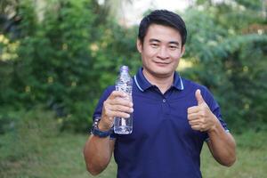 asiático hombre sostiene Bebiendo agua botella, pulgares arriba. concepto, bebida agua para bueno salud. alivio desde sediento, y proteger desde calor ataque, frio abajo en caliente clima condición. salud cuidado. foto