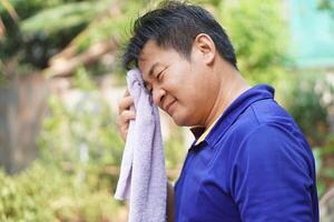 Asian man feel headache and unwell from hot weather condition, use cool wet cloth to wipe face for relief symptom. Concept, self first aid to protect from heat stroke, cool down. Health care. photo