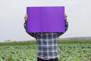 Farmer hold blank violet paper poster at garden background. Concept, Agriculture occupation. Copy space for adding text or advertisement. Protest. Announcement. photo