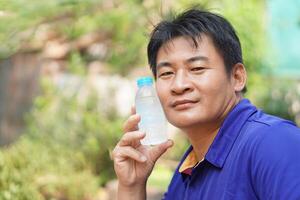 Handsome Asian man hold cool drinking water bottle to touch his cheek for relief hot weather condition. Concept, self first aid to protect from heat stroke, cool down. Health care. photo