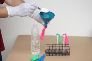 Closeup hands wears gloves holds funnel and flat pink balloon, put spoon of baking soda powder to pour into balloon. Concept, science experiment about reaction of chemical substance . photo