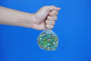Close up hand hold net bag of marbles balls, small spherical object often made from glass. Blue background. Concept. object use as toy for playing games or use for DIY craft decoration. photo