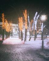 invierno parque a noche con Navidad decoraciones, brillante linternas, pavimento cubierto con nieve y arboles Clásico película estético. foto