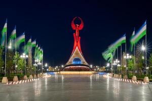 uzbekistán, Tashkent - mayo 5, 2023 iluminado Monumento de independencia en el formar de un estela con un humo pájaro, fuentes y ondulación banderas en el nuevo Uzbekistán parque a Noche. foto