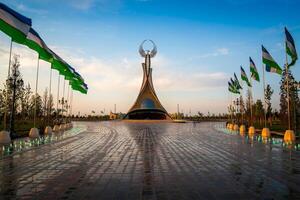 UZBEKISTAN, TASHKENT - MAY 5, 2023 Fountains in a background of monument of Independence in the form of a stele with a Humo bird and waving flags on a sunset after rain in the New Uzbekistan park. photo
