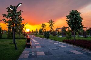 City park in early summer or spring with pavement, lanterns, young green lawn, trees and dramatic cloudy sky on a sunset or sunrise. photo