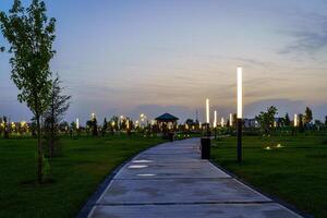 City night park in early summer or spring with pavement, lanterns, young green lawn and trees. photo