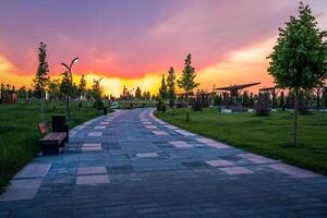 City park in early summer or spring with pavement, lanterns, young green lawn, trees and dramatic cloudy sky on a sunset or sunrise. photo