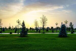 City park in early summer or spring with lanterns, young green lawn, trees and dramatic cloudy sky on a sunset or sunrise. photo