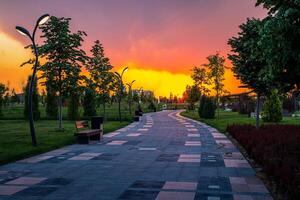 City park in early summer or spring with pavement, lanterns, young green lawn, trees and dramatic cloudy sky on a sunset or sunrise. photo